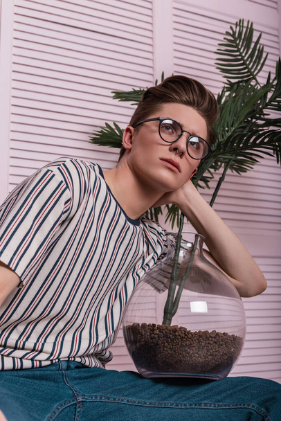 Μοντέλο ενός όμορφου νεαρού άνδρα με κομψά ρούχα σε μοντέρνα γυαλιά με ένα καταπληκτικό λουλούδι σε μια γυάλινη κατσαρόλα που ποζάρει σε μια καρέκλα κοντά σε ένα ροζ ξύλινο τοίχο στο δωμάτιο. Σύγχρονη hipster τύπος χαλαρώνει σε εσωτερικούς χώρους. - Φωτογραφία, εικόνα