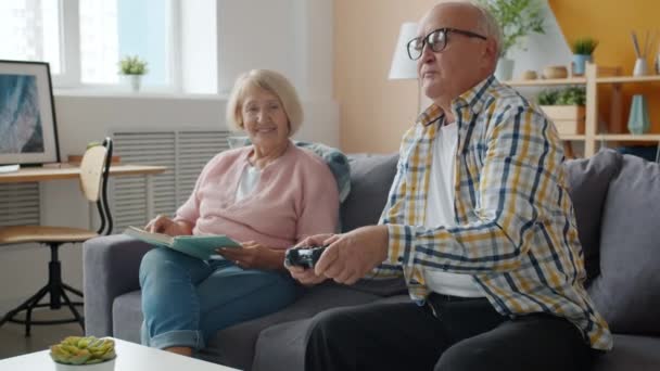 Woman reading book while elderly man playing video game at home on couch - Video