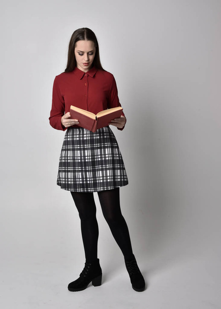 full length portrait of a pretty brunette girl wearing a red shirt and plaid skirt with leggings and boots. Standing pose holding a book, on a  studio background. - Foto, Imagen