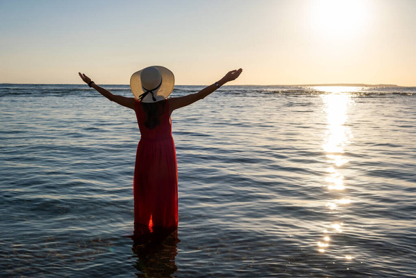 Fiatal nő hosszú piros ruha és szalma kalap felemeli a kezét álló tengervíz a strandon élvezi a kilátás a felkelő nap kora nyáron reggel. - Fotó, kép