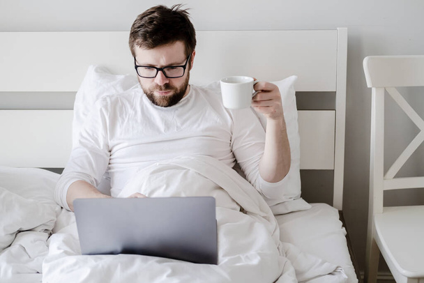 Focused bearded man, with a cup of coffee in his hand, works on a laptop, sitting in bed, under natural light. Modern lifesty. - Fotoğraf, Görsel