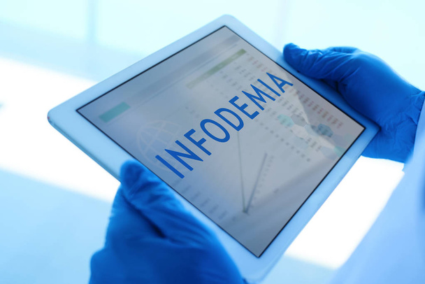 Nahaufnahme eines Arztes, der blaue OP-Handschuhe trägt und ein digitales Tablet in der Hand hält, auf dessen Bildschirm das in Spanisch geschriebene Wort infodemia steht. - Foto, Bild