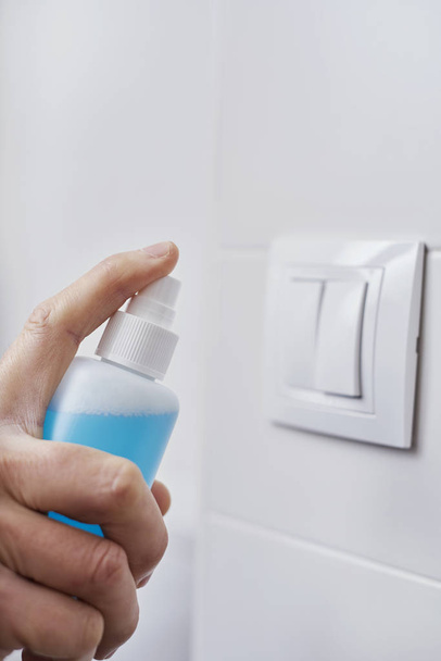 közelkép egy fehér férfi fertőtleníti a villanykapcsolót permetezéssel egy kék fertőtlenítő egy palackból - Fotó, kép