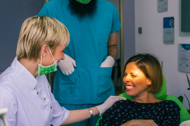 Primer plano retrato de una hermosa joven sentada en la silla dental mientras que el estomatólogo (dentista) con las manos en guantes estériles está arreglando sus dientes. - Foto, imagen