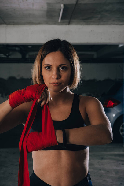 Closeup portrait of a beautiful woman doing red boxing bandages in a garage. - Photo, Image
