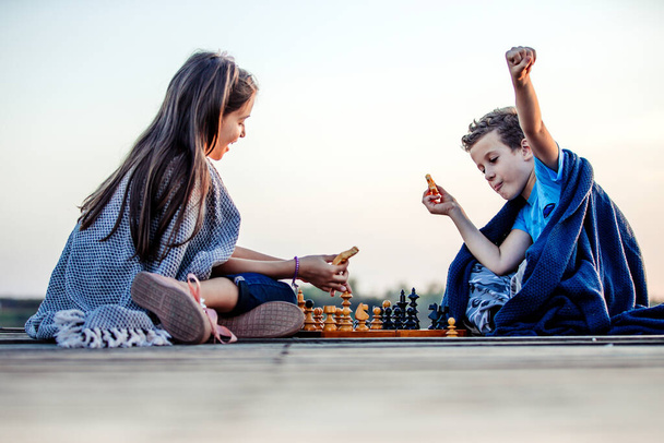 Two young cute little friends, boy and girl having fun while playing chess sitting by the lake in the evening. Kids are playing.Winner gesture. Friendship. - Foto, Imagem