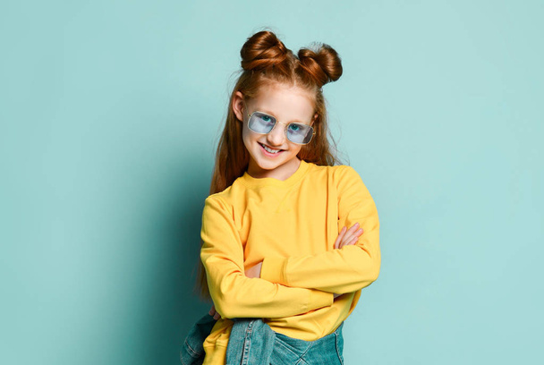 Nice smiling red-haired teen girl in blue glasses and yellow sweatshirt is standing with her arms crossed at chest - 写真・画像