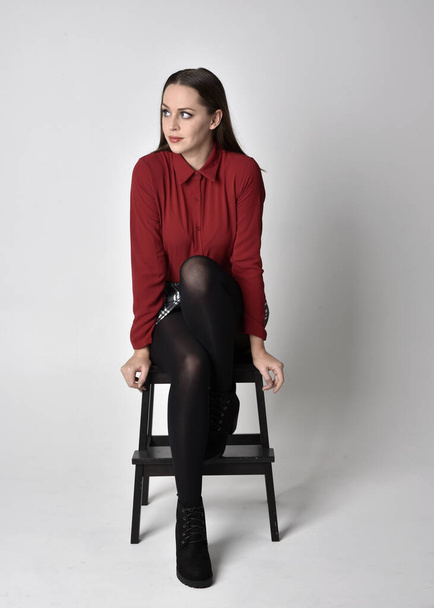 teljes hosszúságú portré egy csinos barna lányról piros ingben és kockás szoknyában, nadrággal és csizmával. Ül egy széken szemben a stúdió háttér. - Fotó, kép