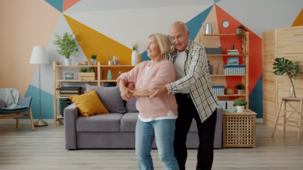 Smiling elderly lady and man dancing at home holding hands enjoying music indoors - Filmagem, Vídeo