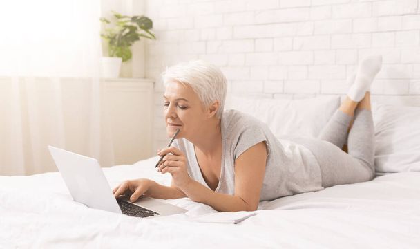 Pensive senior dame browsen op laptop in bed - Foto, afbeelding
