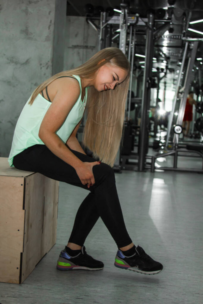 A woman with long blond hair feels severe knee pain during a workout in the gym. People, fitness and healthcare concept. It hurts, is dangerous, anxiety, muscle strain, contusion - Valokuva, kuva