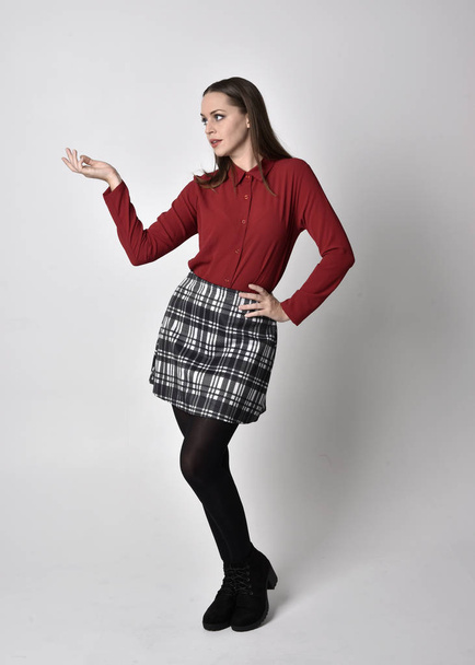 full length portrait of a pretty brunette girl wearing a red shirt and plaid skirt with leggings and boots. Standing pose with hand gesture against a  studio background. - Photo, Image