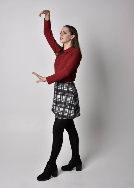 full length portrait of a pretty brunette girl wearing a red shirt and plaid skirt with leggings and boots. Standing pose with hand gesture against a  studio background. - Photo, Image