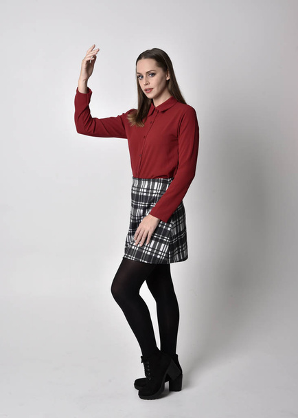 full length portrait of a pretty brunette girl wearing a red shirt and plaid skirt with leggings and boots. Standing pose with hand gesture against a  studio background. - Fotografie, Obrázek