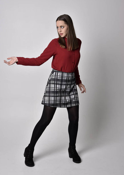 full length portret van een mooi brunette meisje met een rood shirt en geruite rok met legging en laarzen. Staande pose met de hand gebaar tegen een studio achtergrond. - Foto, afbeelding