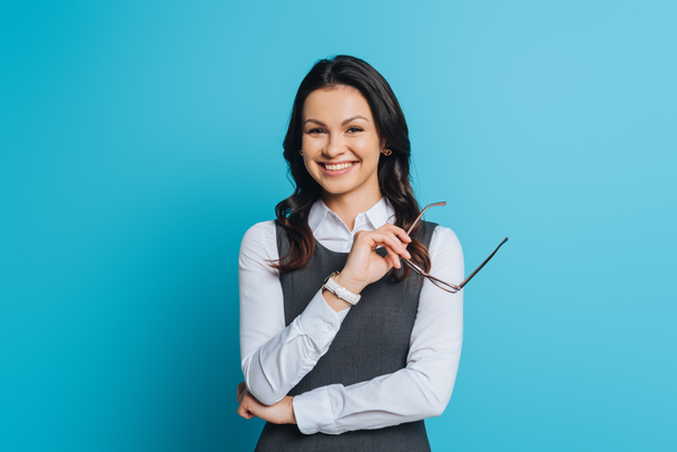 cheerful businesswoman holding eyeglasses while smiling at camera on blue background - Photo, Image