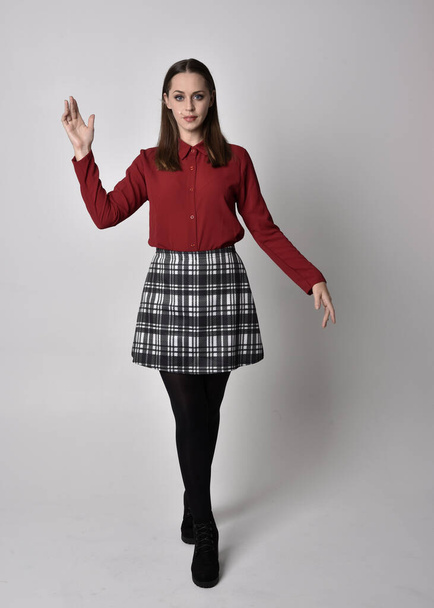 full length portrait of a pretty brunette girl wearing a red shirt and plaid skirt with leggings and boots. Standing pose with hand gesture against a  studio background. - Φωτογραφία, εικόνα