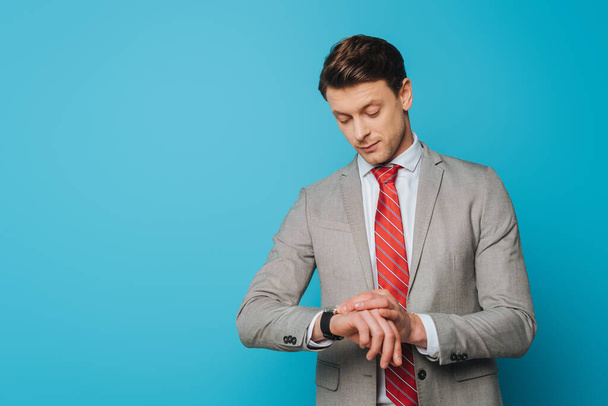 handsome, serious businessman checking time on wristwatch on blue background - Zdjęcie, obraz