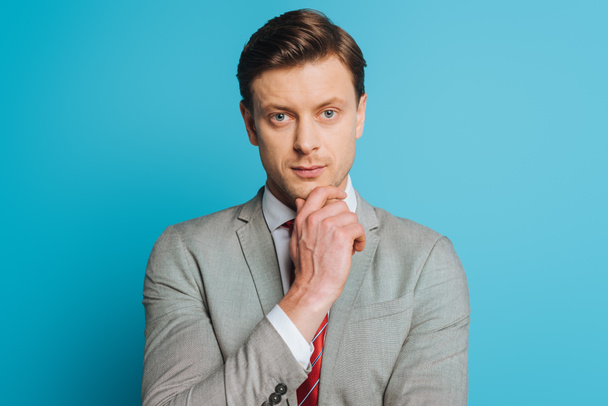 serious, thoughtful businessman touching chin while looking at camera on blue background - Фото, зображення