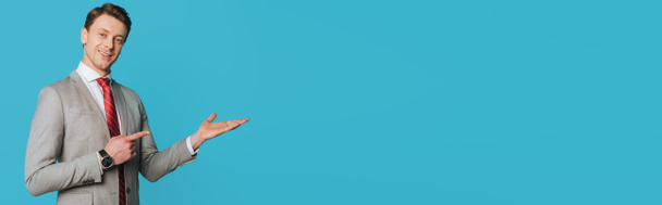 plano panorámico de un hombre de negocios sonriente señalando con el dedo mientras está de pie con el brazo abierto y mirando a la cámara aislada en azul
 - Foto, Imagen