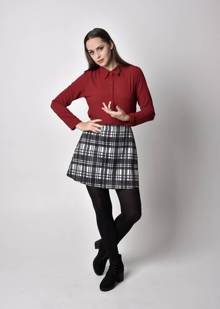 full length portrait of a pretty brunette girl wearing a red shirt and plaid skirt with leggings and boots. Standing pose with hand gesture against a  studio background. - Photo, image