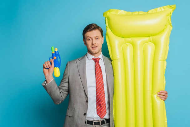 smiling businessman holding inflatable mattress and water gun on blue background - Foto, Imagen