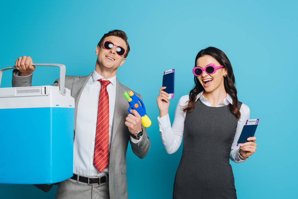 excited businesswoman with passports and air tickets near businessman holding portable fridge and water gun on blue background - Φωτογραφία, εικόνα