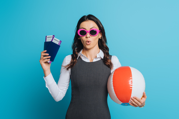 verrast zakenvrouw in zonnebril met opblaasbare bal, paspoorten en vliegtickets op blauwe achtergrond - Foto, afbeelding
