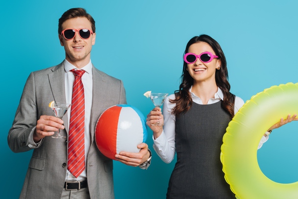 two cheerful businesspeople with glasses of cocktail, inflatable ball and swim ring looking at camera on blue background - Photo, Image