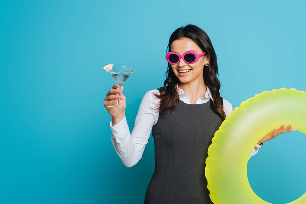 wesoła bizneswoman w okularach przeciwsłonecznych trzymając szklankę koktajlu i pierścień pływacki na niebieskim tle - Zdjęcie, obraz