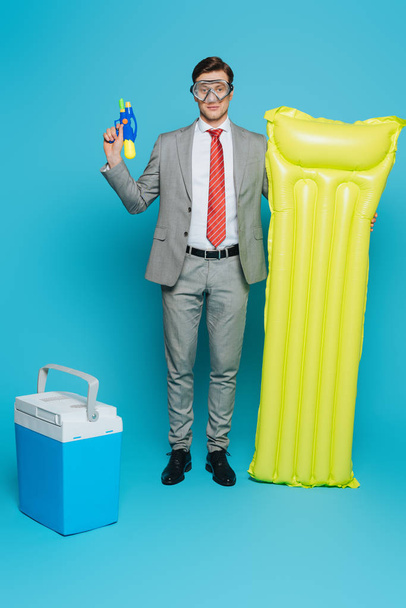 homem de negócios novo na máscara de mergulho que prende o colchão inflável e a arma da água ao estar perto da geladeira portátil no fundo azul
 - Foto, Imagem