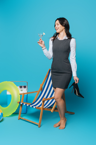 happy businesswoman holding high heeled shoes and glass of cocktail near deck chair, portable fridge and inflatable ring on blue background - Photo, Image