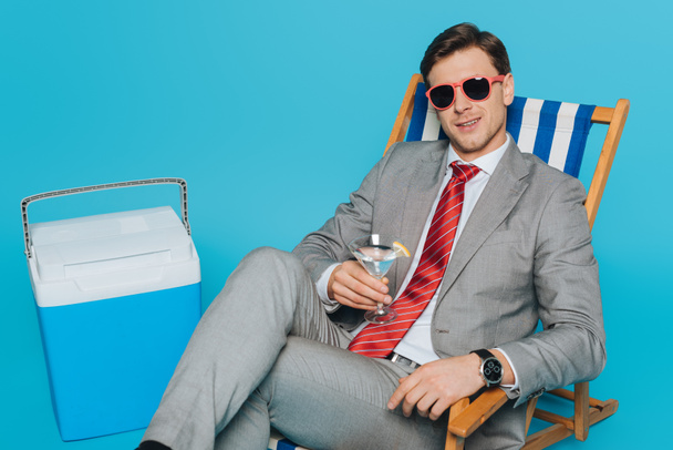 homem de negócios feliz em óculos de sol sentado na cadeira deck com copo de coquetel perto da geladeira portátil no fundo azul
 - Foto, Imagem