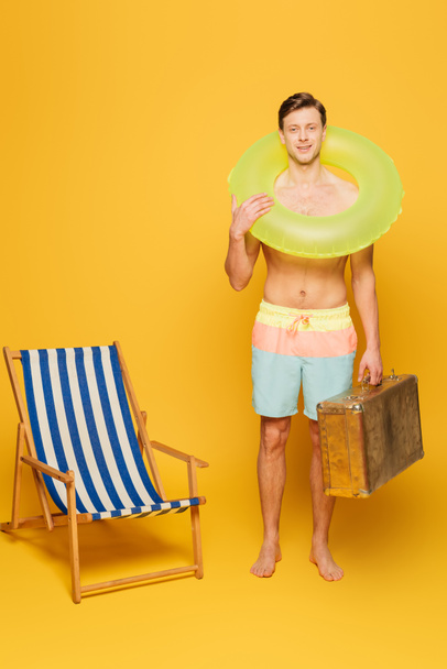 shirtless man in shorts with vintage suitcase and swim ring standing near deck chair on yellow background - 写真・画像