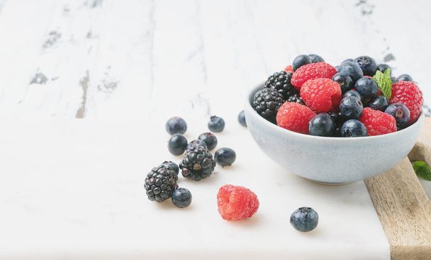 Ripe and juicy fresh berries in the bowl and on the marble wooden board over white rustic wooden backgrounds. Blueberry, Raspberry and Blackberry. Side view. Selective focus. - Photo, Image