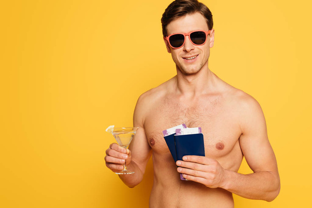 shirtless man in sunglasses holding glass of cocktail, passports and air tickets on yellow background - Photo, image
