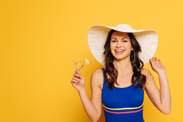 улыбающаяся женщина в синем купальнике и шляпе, держащая стакан коктейля и смотрящая на камеру, изолированную на желтом
 - Фото, изображение