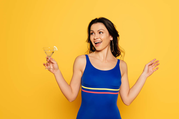 veselá žena v modrých plavkách drží sklenici koktejlu a dívá se stranou izolované na žluté - Fotografie, Obrázek