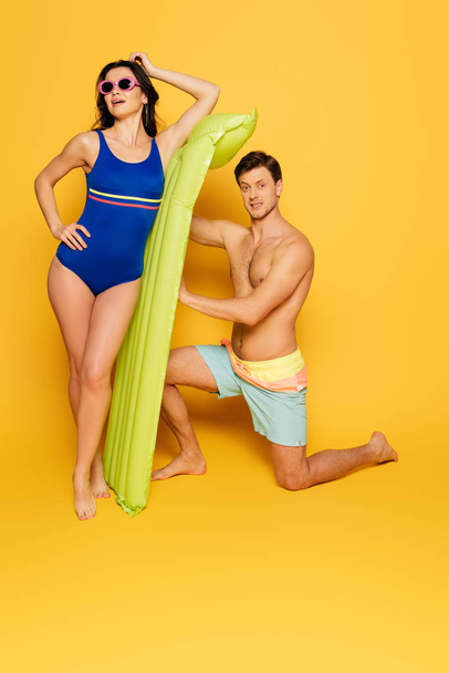 attractive woman posing in sunglasses and shirtless man standing on knee near inflatable mattress on yellow background - Photo, Image
