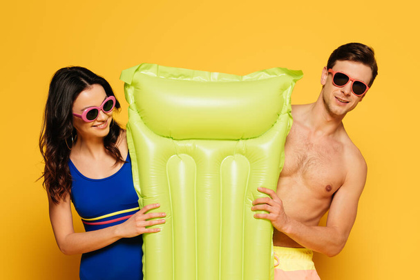 cheerful couple in sunglasses holding inflatable mattress on yellow background - Valokuva, kuva
