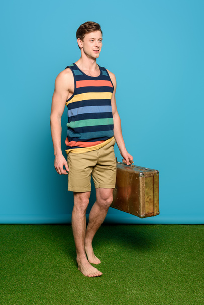 handsome man in striped singlet and shorts holding vintage suitcase on blue background - Foto, imagen
