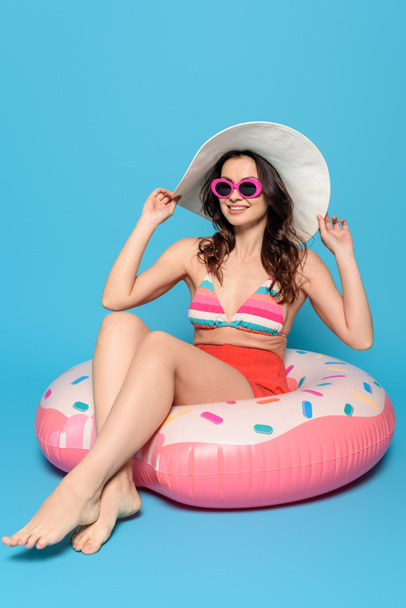 cheerful, stylish woman in sunglasses touching sun hat while sitting on swim ring on blue background - Fotoğraf, Görsel
