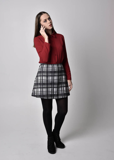 full length portrait of a pretty brunette girl wearing a red shirt and plaid skirt with leggings and boots. Standing pose holding smartphone, against a studio background. - Fotoğraf, Görsel