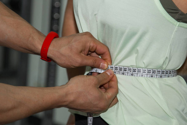 weight loss, personal trainer, nutritionist, fitness, motivation, healthy lifestyle. A man measures the waist of a girl with a tape measure. Close up, macro photo. Preparation for training - Valokuva, kuva