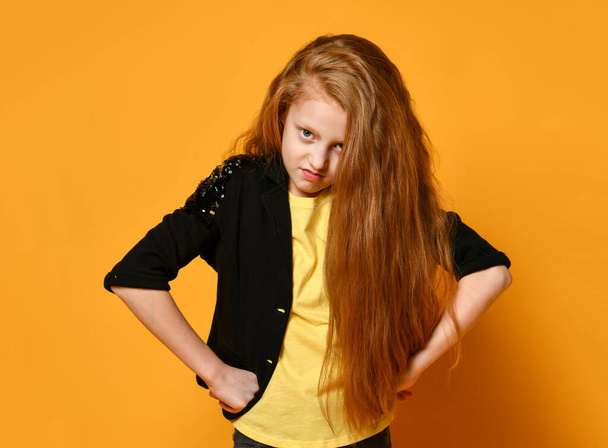 Miúdo adolescente de casaco preto e t-shirt amarela. Ela está chateada, com as mãos nos quadris, posando no fundo laranja. Fechar
 - Foto, Imagem