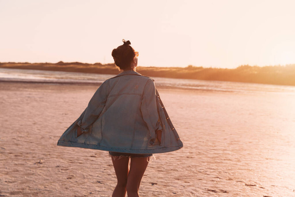 Young stylish hipster girl dancing Jeans jacket Sunset coast during travel summer holidays vacation outdoors - 写真・画像