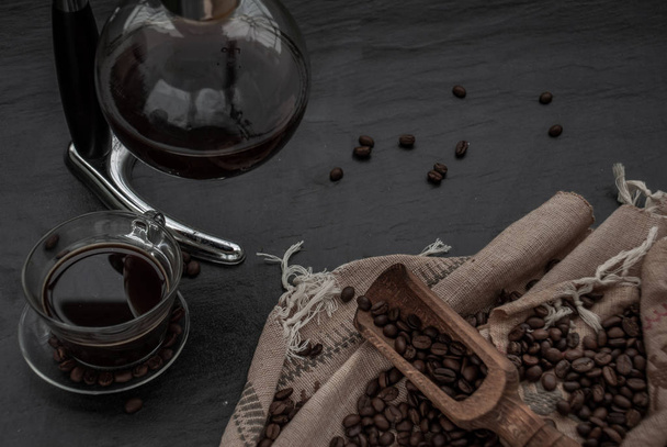 Kávový styl, šálek kávy a kávové boby, které se rozsytá starými dřevěnými naběračkami a kávovou fazolí na dřevěném a tmavém kamenném pozadí. Nakloněné zobrazení shora s místem kopírování textu. - Fotografie, Obrázek