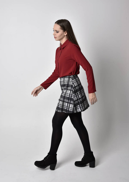 full length portrait of a pretty brunette girl wearing a red shirt and plaid skirt with leggings and boots. Standing pose in side profile, against a studio background. - Photo, Image