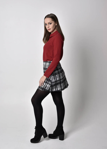 full length portrait of a pretty brunette girl wearing a red shirt and plaid skirt with leggings and boots. Standing pose in side profile, against a studio background. - Photo, Image