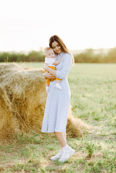 eine junge hübsche Mutter mit einem kleinen Kind auf dem Arm steht in einem Kleid inmitten eines Feldes in der Nähe eines gesammelten Heuhaufens in den Strahlen des goldenen Sonnenuntergangs - Foto, Bild